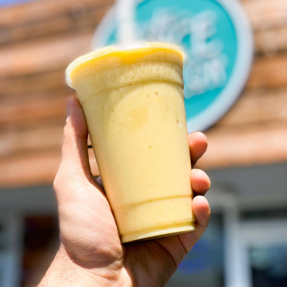 A hand holds a yellow smoothie in a clear plastic cup, with the Juice Parlor NoHo logo blurred in the background.