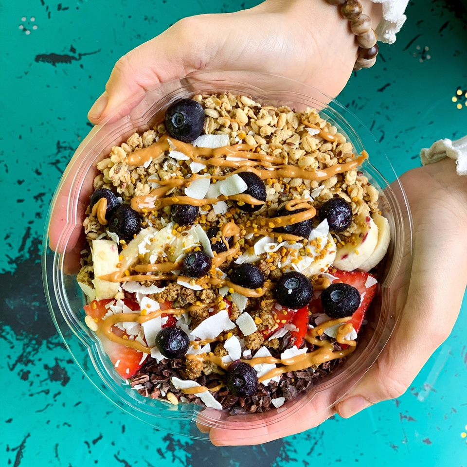 Hands holding a Juice Parlor NoHo acai bowl with granola, blueberries, strawberries, bananas, and peanut butter drizzle.