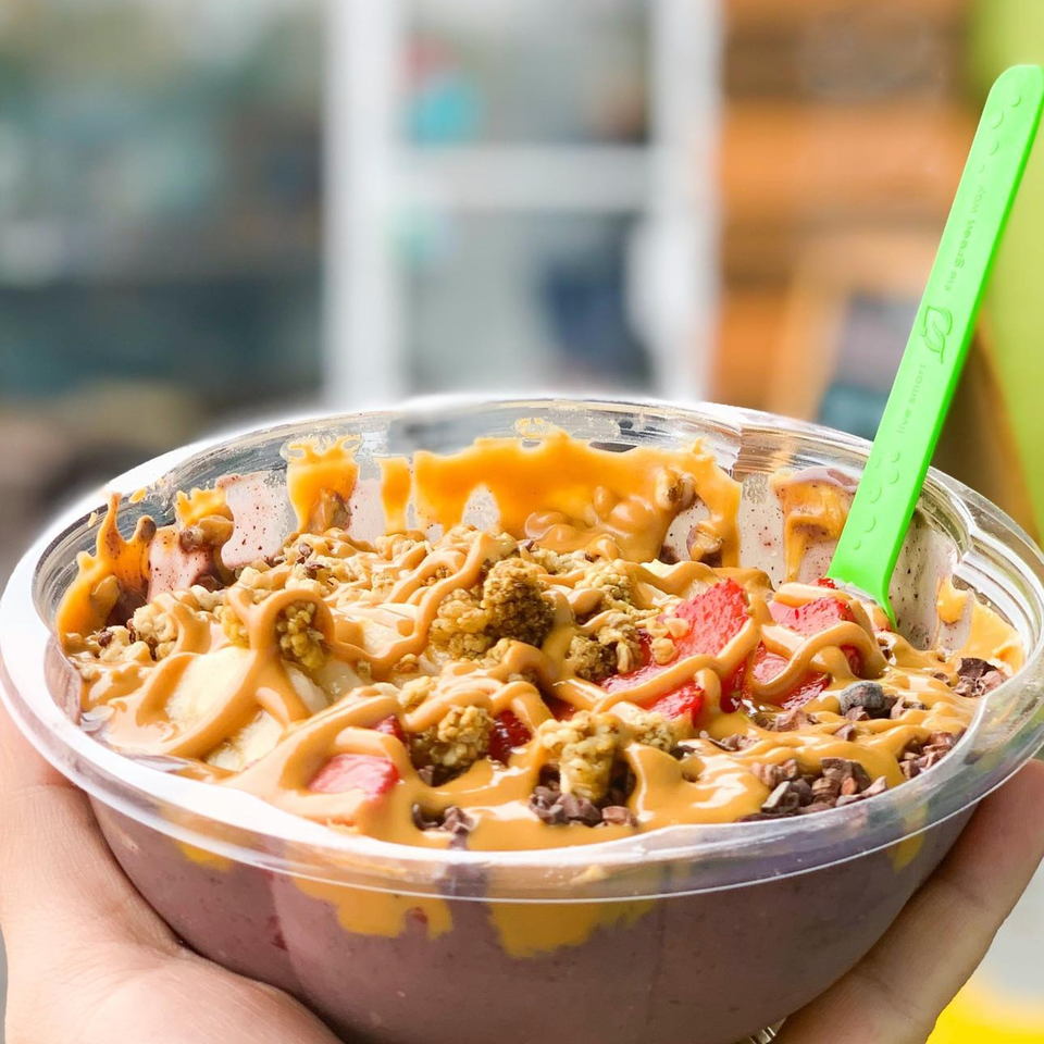 A hand holds a Juice Parlor NoHo acai bowl topped with peanut butter, granola, and strawberries.